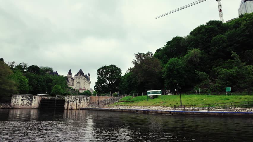 Cinematic boat-level view of the historic Rideau Canal Locks and the iconic Château Laurier hotel in Ottawa, Ontario, Canada. Captured under dramatic overcast skies with lush green surroundings