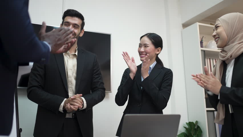 A businessman shakes hands with a colleague while diverse teammates applaud their shared business success.