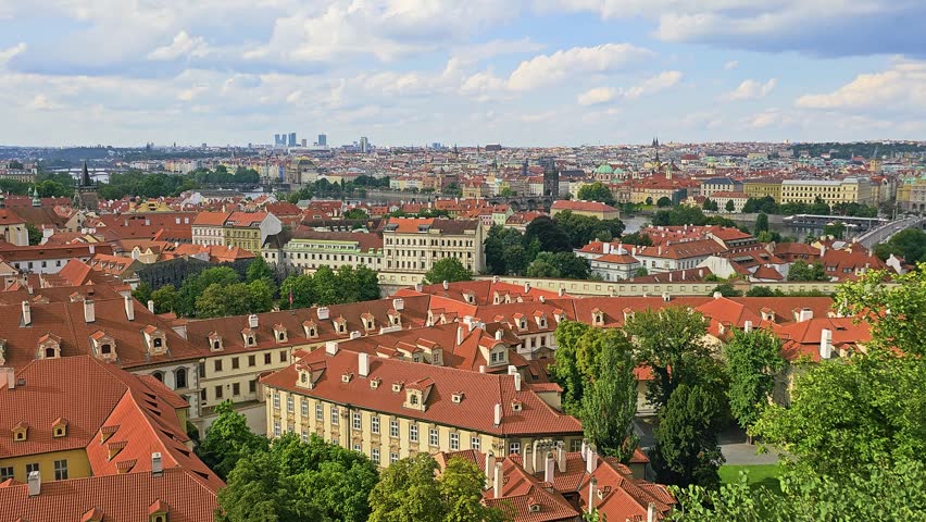 Elevated travel view of Prague, Czech Republic, featuring historic red rooftops, palace courtyards, Vltava River, old bridges, church spires, and layered urban skyline under clouds.