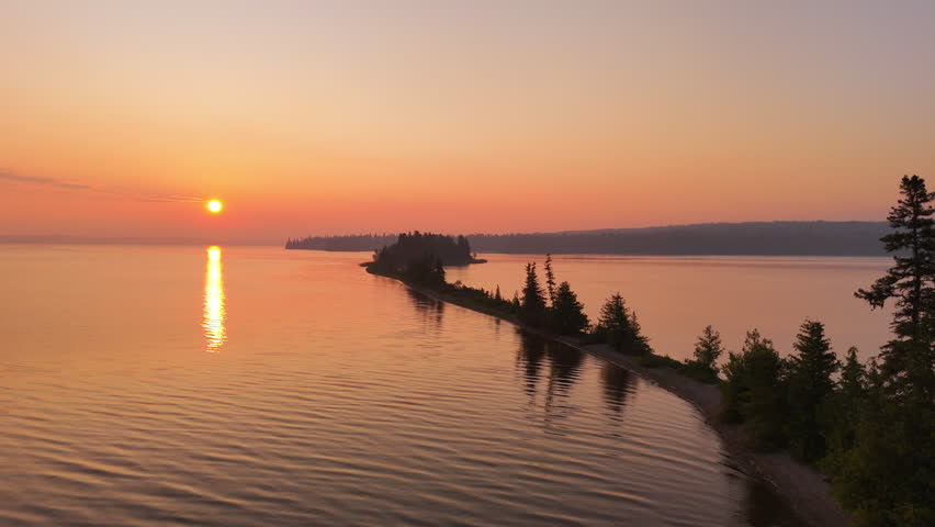 Drone glides toward a narrow sandy peninsula surrounded by calm water beneath a vivid orange sunrise sky in Nagagamisis Provincial Park, Northern Ontario.