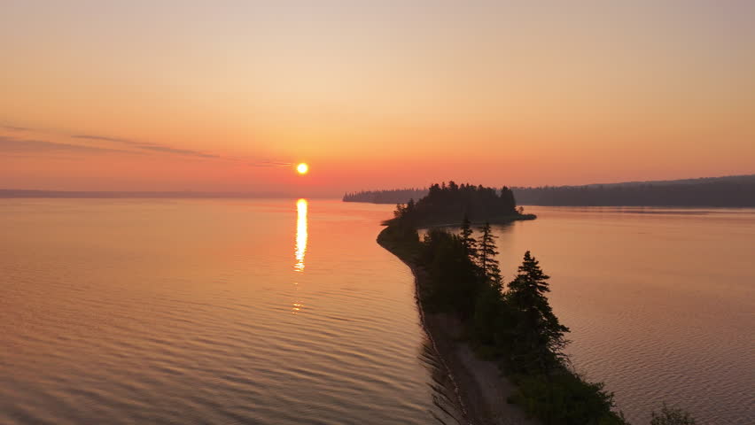Aerial view follows a narrow sandy peninsula surrounded by calm water under a warm sunrise sky softened by distant wildfire smoke in Nagagamisis Provincial Park.