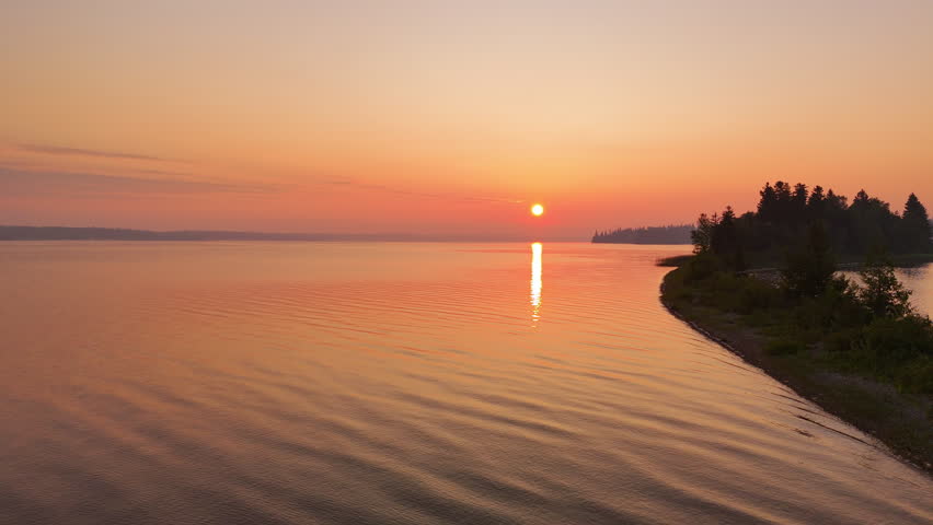 Drone pulls back to reveal a narrow sandy peninsula extending into calm water beneath a glowing orange sunrise sky in Nagagamisis Provincial Park.
