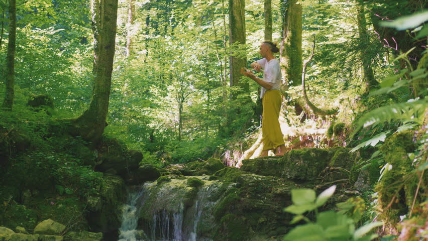 A person balances on moss-covered rocks beside a small waterfall in a lush green forest, practicing zen-like yoga to embody a healthy lifestyle and relaxation.