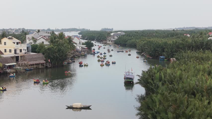 Coconut-shaped boats filled with people travel down a calm river lined with dense palm trees and riverside houses in Hoi An, Vietnam.