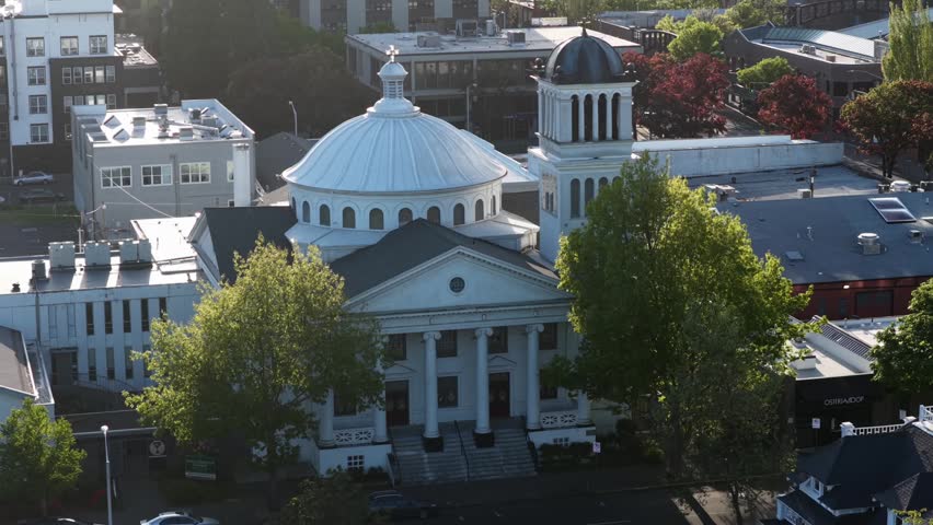 Historic domed church with white columns anchors downtown Eugene, Oregon at golden hour, surrounded by leafy green trees plus mid-rise buildings under warm evening light. Aerial drone wide shot