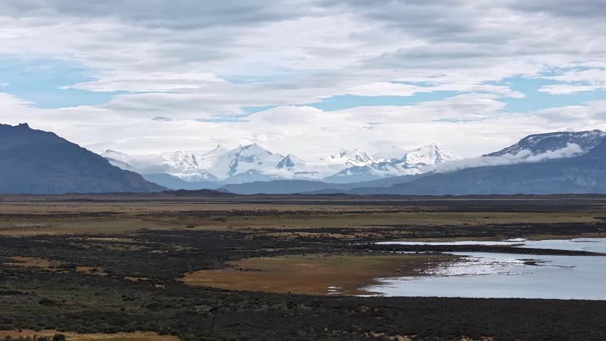 Panoramic view of the Andes mountains in Patagonia, Argentina, with snow capped peaks, vast plains, and a lake under a cloudy sky