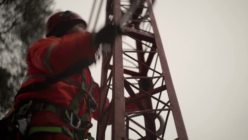 A Middle Eastern worker in a vest and helmet, ascending a tower in the construction site, with gray sky in the background