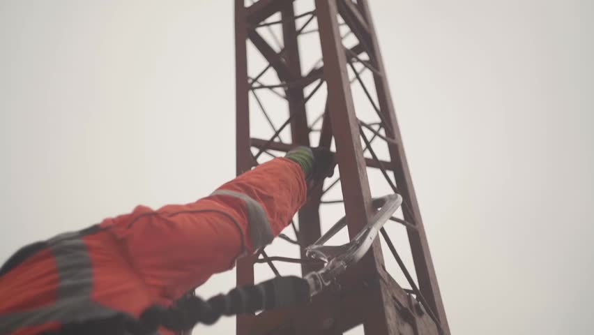 A Middle Eastern worker in a vest and helmet, ascending a tower in the construction site, under gray sky