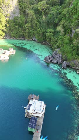 Stunning high-angle aerial view of a solar-powered floating houseboat anchored in clear turquoise lagoon water surrounded by lush jungle cliffs and a paddleboard in Palawan, Philippines.