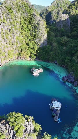Stunning high-angle aerial view of a solar-powered floating houseboat anchored in clear turquoise lagoon water surrounded by lush jungle cliffs and a paddleboard in Palawan, Philippines.