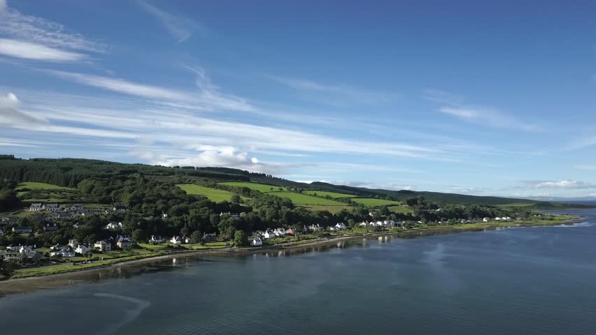 An aerial view captures Isle of Arran, long coastline with a town nestled between green hills and a body of water under a blue sky in Scotland