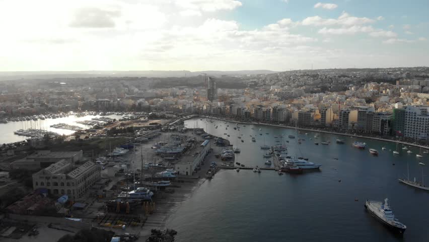 A drone scene of Grand Harbor with boats and waterfront buildings in Valletta under a cloudy sky, Malta