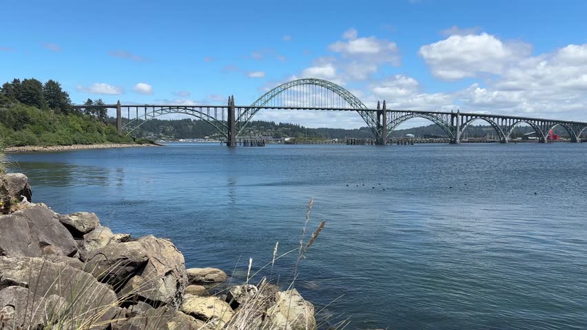 The historic steel arch Yaquina Bay Bridge stretches across calm harbor waters, viewed from a rocky shoreline in Newport, Oregon.