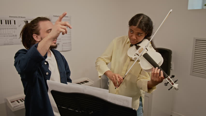 Young Caucasian man teaching African American teenage girl to play violin during music lesson. Ideal for promoting private tutoring, music schools, and creative arts education