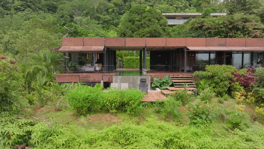 Aerial view of a luxurious modern villa in Costa Rica featuring an infinity pool and wooden deck with lounge chairs nestled among lush tropical rainforest vegetation on a misty hillside.