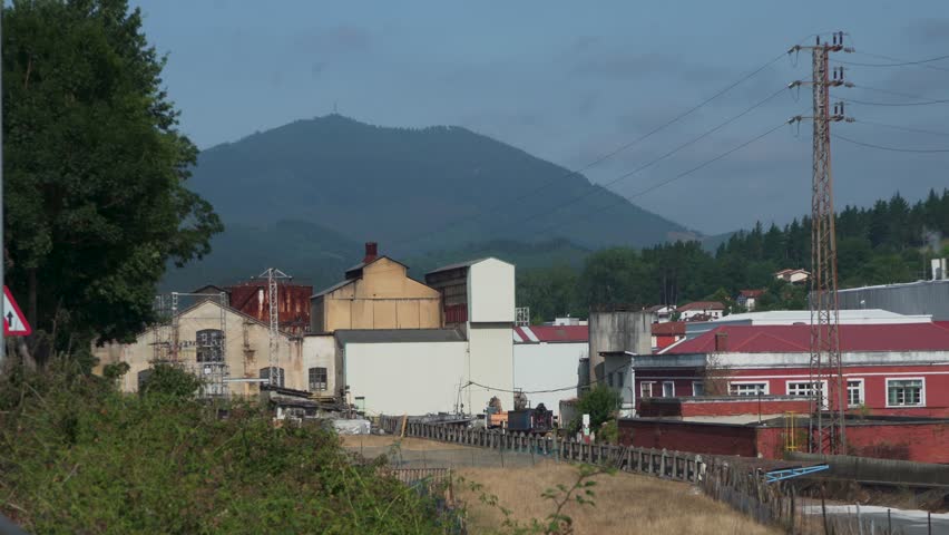 Editorial wide shot of an industrial factory featuring a water tower, production buildings, conveyor systems, and manufacturing infrastructure in the Basque Country, Spain.
