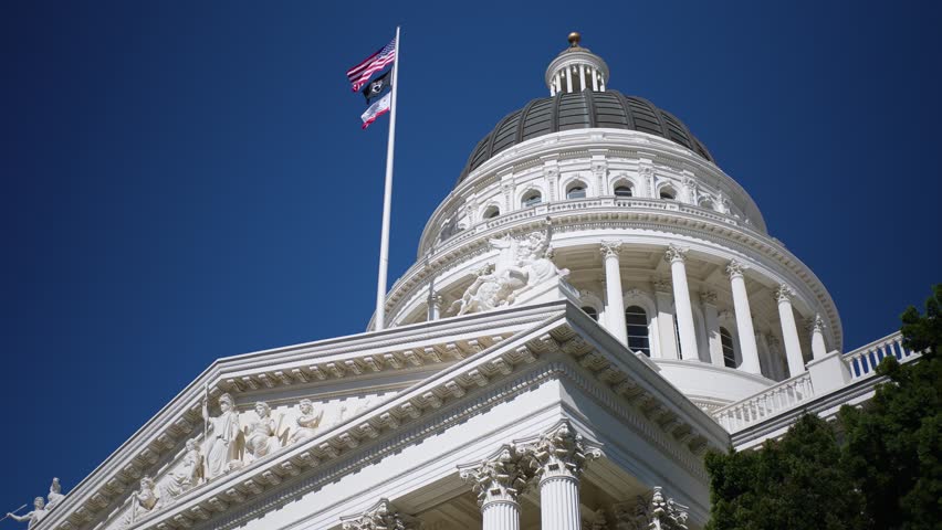 The California State Capitol stands tall under bright sun in Sacramento with the flag proudly flying from the dome.