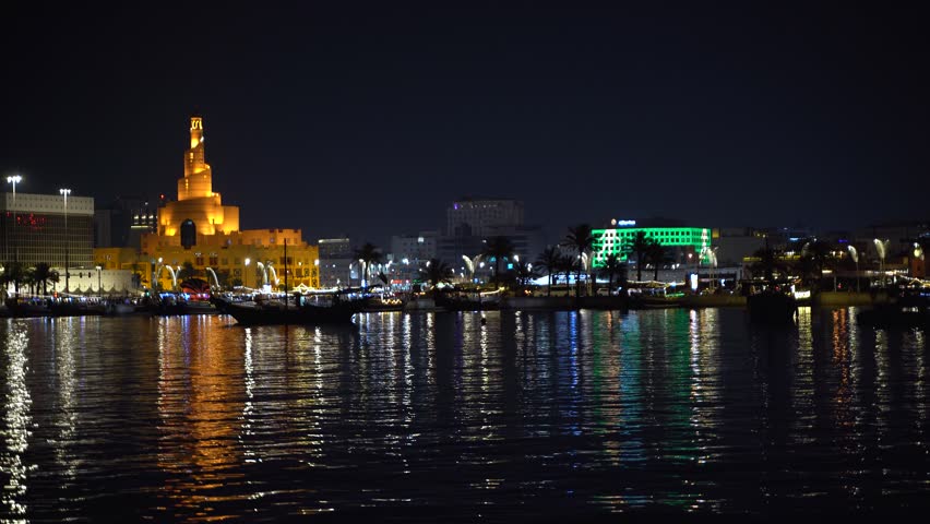 Doha corniche at night with traditional dhow boats moored along the waterfront, the illuminated FANAR spiral minaret, palm trees, and city buildings reflecting on calm water, Qatar