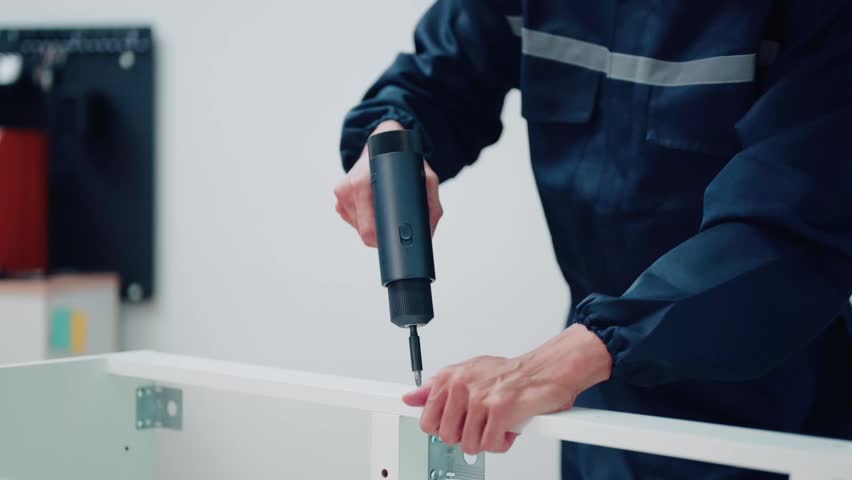 A worker in a navy blue uniform assembles white furniture using a cordless electric screwdriver, ideal for home improvement and repair service concepts.