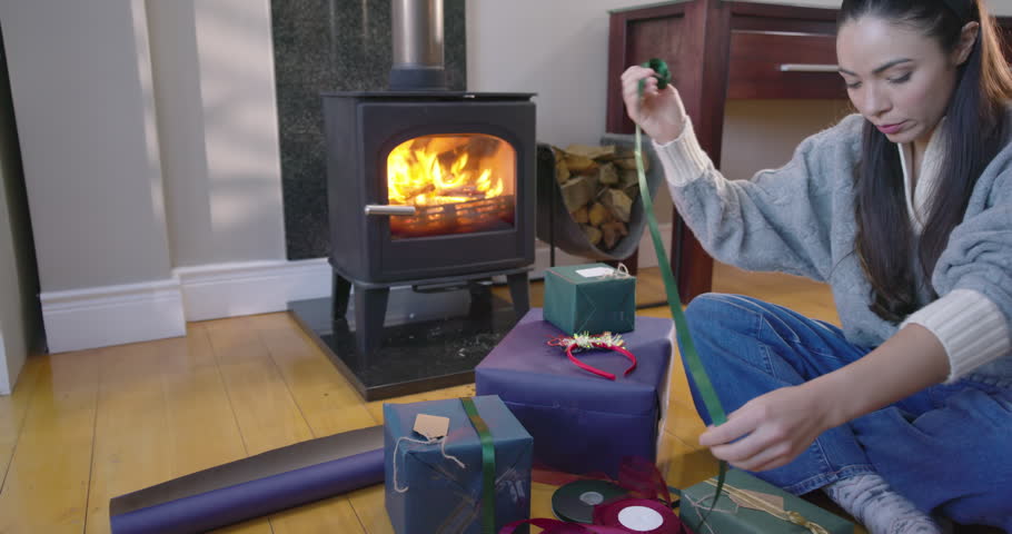 Asian woman sitting by wood stove, preparing gifts, wrapping with ribbons and twine, copy space. Cozy, hearth, fireplace, presents, packaging, scissors, craft