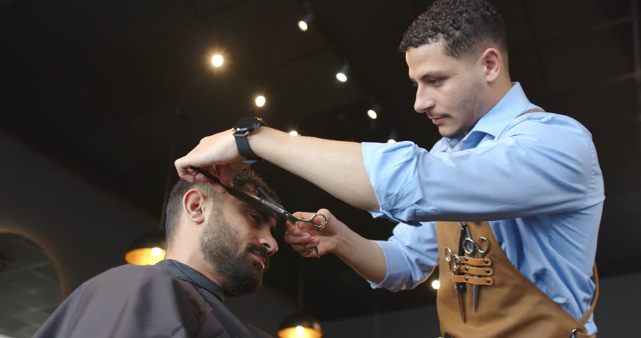 Barber trimming client, lifting hair with comb and scissors to finish haircut at shop, copy space. Grooming, salon, precision, tools, apron, lighting, closeup