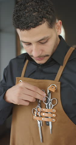 Vertical video: African man barber in apron looking down checking scissors in shop readying tools. Leather, strap, rivets, gear, equipment, hairstylist, vertical video