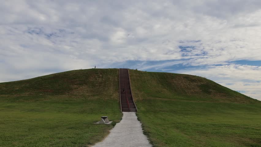 A wide view of a large grassy earthen mound featuring a long central staircase leading to the summit with a few people visible at the top.