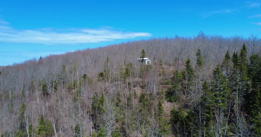 Scenic countryside drone shot featuring a lone white cabin perched above a forested hillside in brilliant daylight.