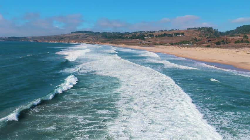 Stunning 4K aerial drone footage of the iconic Big Sur coastline in California, featuring powerful ocean waves rolling onto a long sandy beach with crystal-clear turquoise seawater. Captured under a bright blue sky, this breathtaking coastal landscape showcases the rugged beauty of the Pacific Ocean. Perfect for travel films, tourism promotions, documentaries, nature projects, cinematic backgrounds, and coastal scenery.