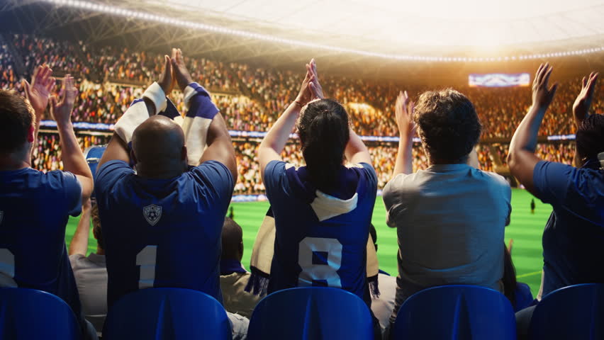 Soccer supporters cheer in packed stadium stands during a football match, creating loud crowd energy and team support.