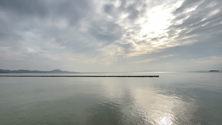 This high-quality 4K footage captures the tranquil beauty of Taihu Lake at the Yuantouzhu (Turtle Head Isle) scenic area in Wuxi, China. The scene features a peaceful water surface reflecting a soft, overcast sky, with distant mountains and a long stone breakwater creating a minimalist and serene atmosphere. This static shot is perfect for travel documentaries, meditation videos, cinematic backgrounds, or corporate projects seeking a calm, professional, and nature-themed aesthetic.