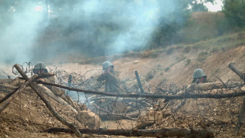 Historical military reenactment showing soldiers taking cover in a trench behind barbed wire as a blast sends smoke and dirt into the air.