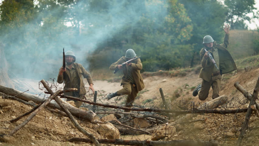 Historical military reenactment showing soldiers running through smoke and debris over a trench under fire.