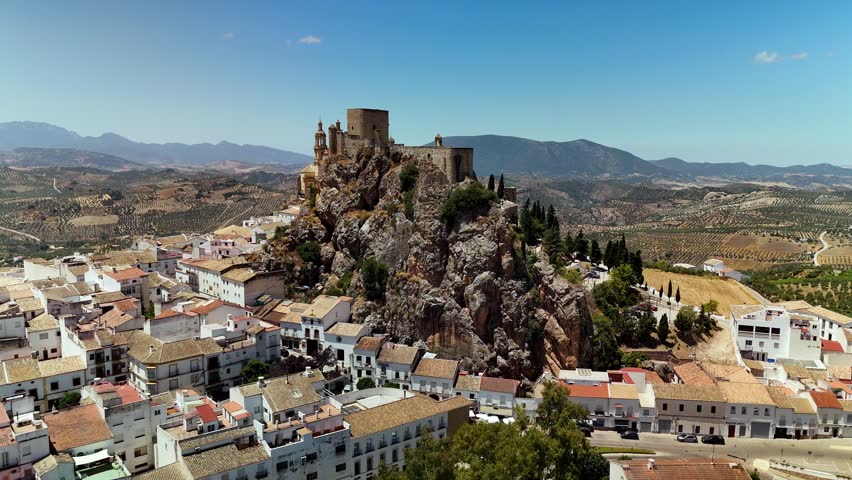 Olvera Castle and the Nuestra Senora de la Encarnacion church on a rocky hilltop in Andalusia, Spain. A low altitude drone orbit reveals the white-washed houses of the pueblo blanco against the summer countryside.