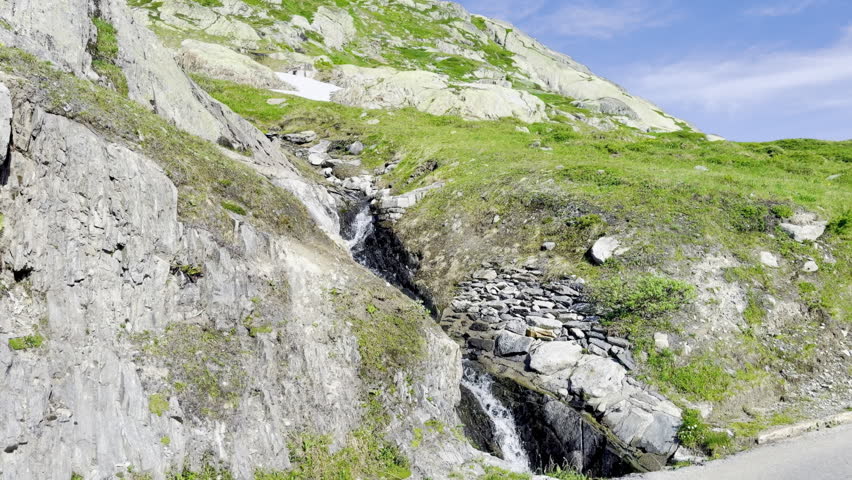 Alpine mountain stream cascading over polished granite slabs and rocks below lingering snow patches on a high hillside near Grimsel Pass in the Bernese Alps of Switzerland on a clear summer day