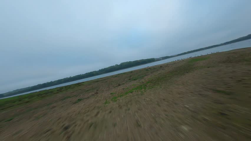 Low angle FPV drone point of view flying fast over a large lake with wild ducks taking off under gloomy autumn skies.