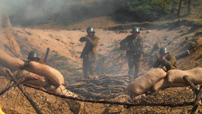 Historical reenactment of soldiers aiming rifles from a smoky trench with sandbags and barbed wire.