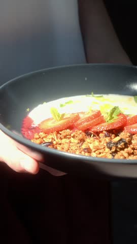 A person holds a breakfast bowl filled with crunchy granola, fresh sliced strawberries, yogurt and mint leaves.