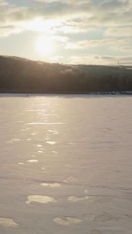 Snowy lake scene at sunset features a solitary person walking on the ice, with warm sunlight reflecting off the surface and trees in the distance