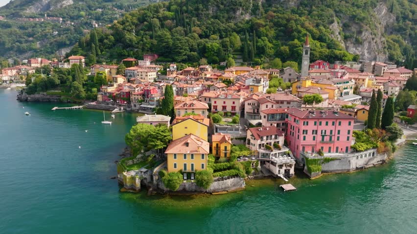 Colorful historic houses sit on steep rocky shores of Lake Como in Varenna, Italy. UHD aerial captures charming waterfront village of Varenna surrounded by calm water and green mountains
