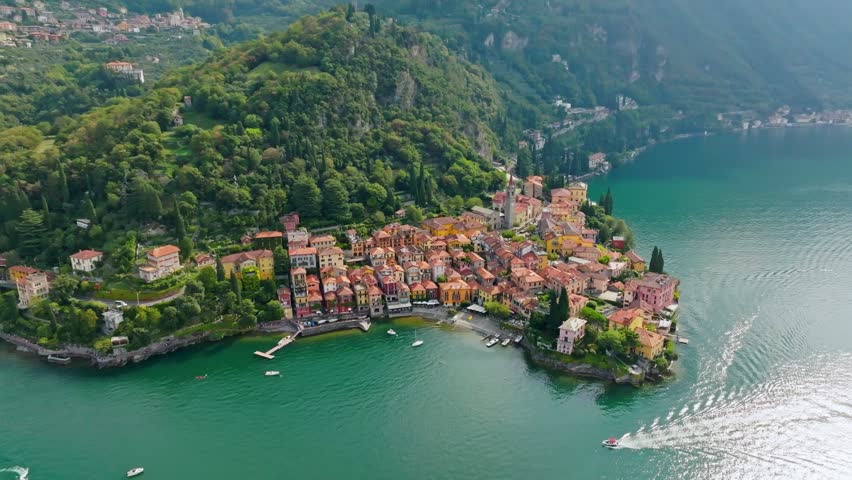 Aerial 4K of beautiful pastel buildings directly on calm blue waters of Lake Como in Varenna, Italy. Historic colorful villas and traditional church tower dominate scenic coastline of Varenna