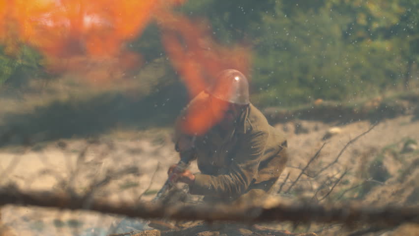 Historical military reenactment showing soldier in a defensive trench position as explosions and flames erupt behind barbed wire.