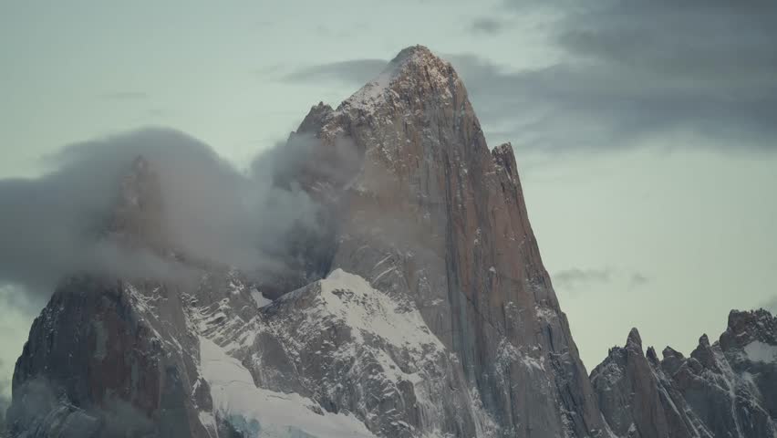 An aerial timelapse of jagged, snow-capped Fitz Roy Mountain range rises dramatically with a cloudy, twilight sky in El Chaoten, Patagonia, Argentina