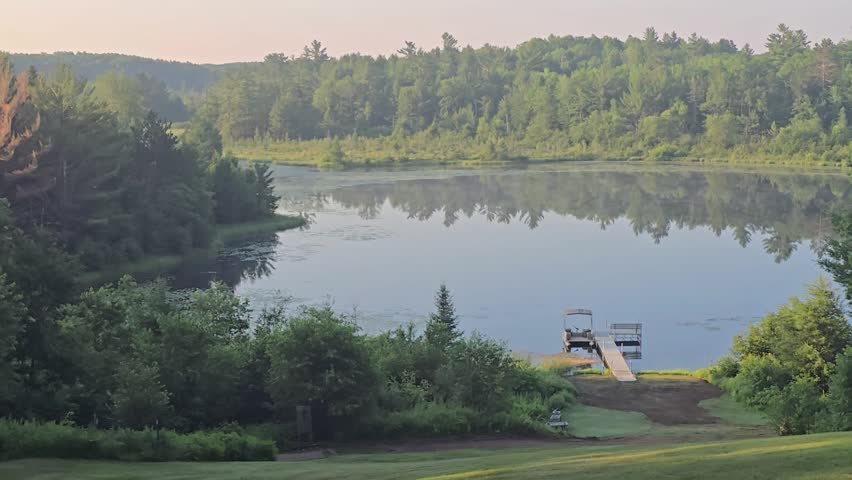 Light morning mist drifts across a calm forest lake with mirror-like reflections and natural birdsong during a peaceful summer sunrise in northern Wisconsin, USA.