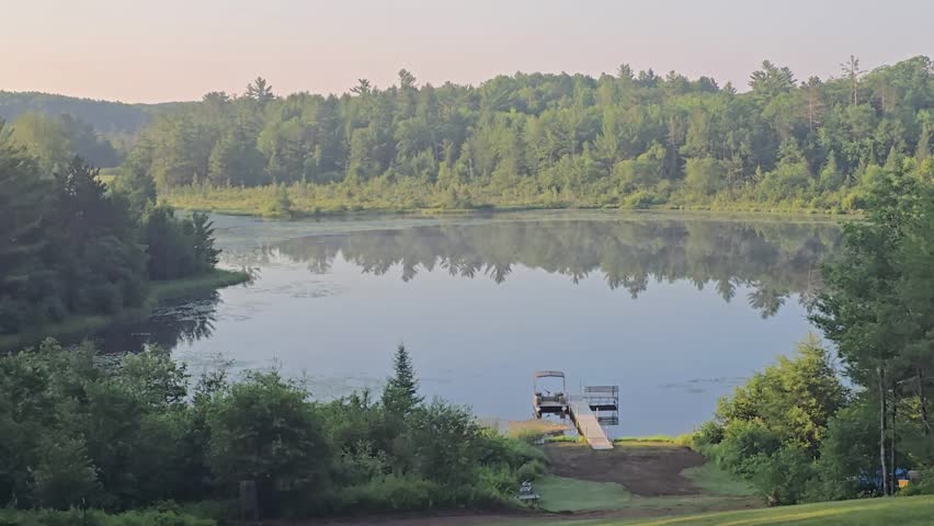 Light morning fog drifts slowly across a calm forest lake with mirror-like reflections as birds sing during a peaceful summer sunrise in northern Wisconsin, USA.