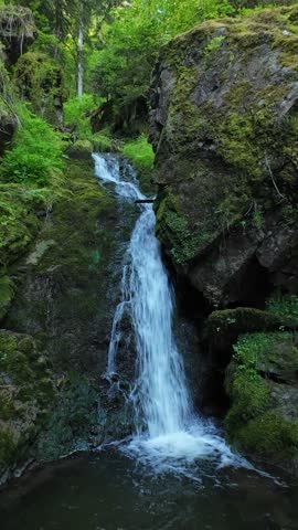 Small natural waterfalls flowing over moss-covered rocks in a lush green forest on a sunny summer day. Peaceful woodland scenery with fresh water and vibrant foliage.