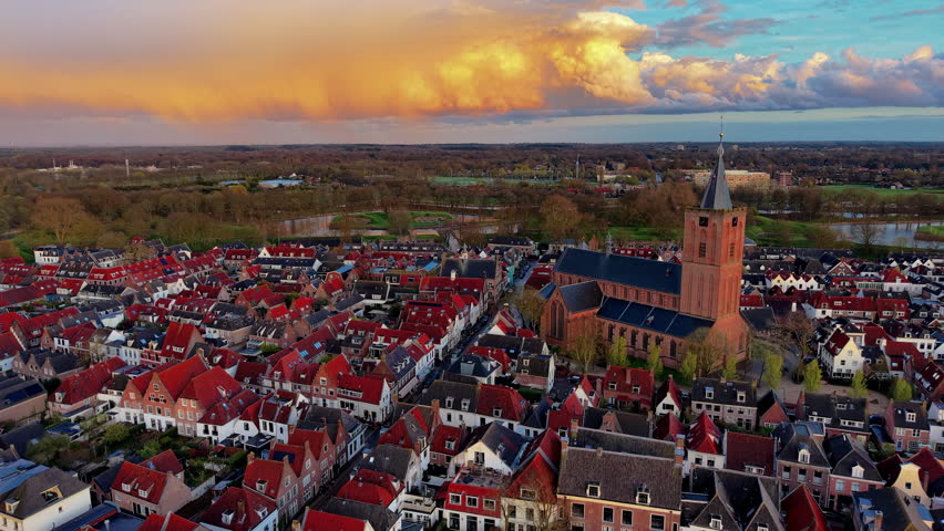 A magnificent wide-angle aerial photograph capturing a breathtaking panoramic sunset view over a historic medieval fortress town. The frame is dominated by a dense grid of traditional European residential houses featuring steep, vibrant red-tiled roofs and classic architecture. Standing prominently on the right is a massive, historic brick church cathedral with a tall, majestic spire pointing toward the heavens. In the background, the town is enclosed by scenic water moats, defensive canals, and lush green park belts with mature trees. The entire landscape is illuminated by the warm, dramatic light of a stunning sunset, with massive storm clouds glowing in vibrant shades of orange, yellow, and purple against the blue sky. This high-resolution capture beautifully documents ancient urban heritage, historical architecture, and dramatic coastal weather patterns.