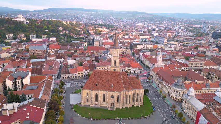 Aerial view of a historic city center with a large church in the foreground, red-roofed buildings, and a hilly urban skyline stretching into the distance. Cluj-napoca in Romania