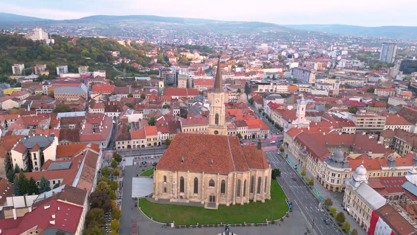 Aerial view of a historic city center with a large church in the foreground, red-roofed buildings, and a hilly urban skyline stretching into the distance. Cluj-napoca in Romania