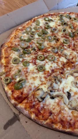 Sliced pizza with melted cheese topped with green olives and mushrooms in cardboard delivery box on wooden table
